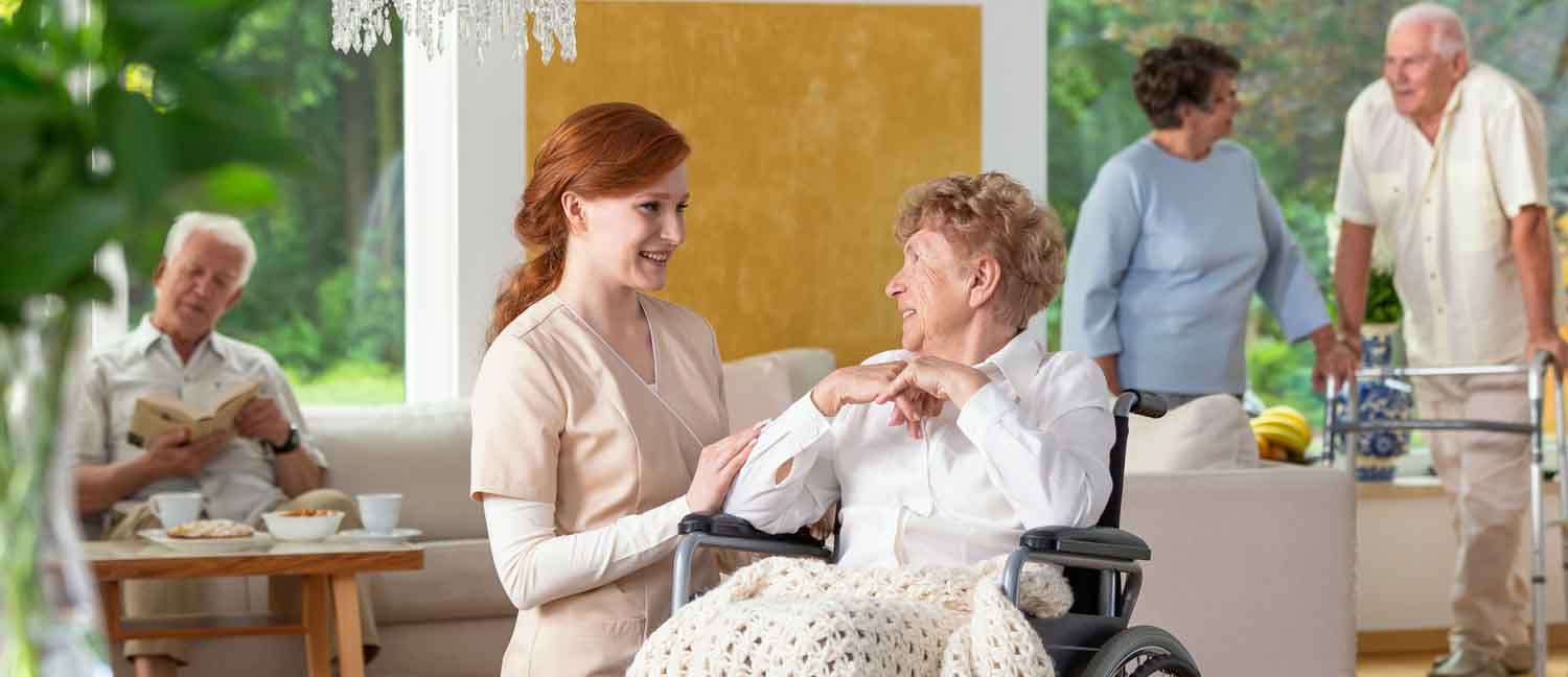 Seniors in a luxury living room of a private retirement home. Tender caregiver by an elderly lady in a wheelchair in the foreground.
